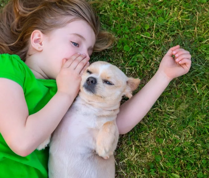 young girl wearing green shirt laying on grass next to her small dog, she is whispering a secret in the dog's ear
